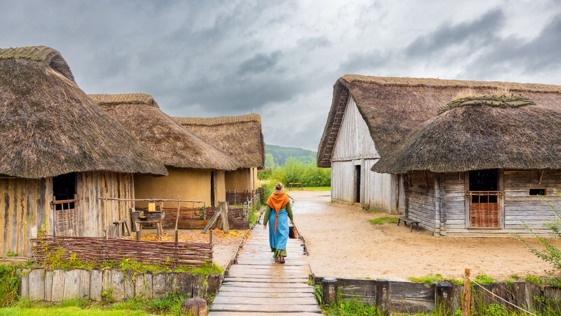 Eine Person in traditioneller Kleidung geht auf einem Holzweg zwischen strohgedeckten Hütten in der UNESCO-Welterbestätte des archäologischen Grenzkomplexes von Haithabu und Danewerk.