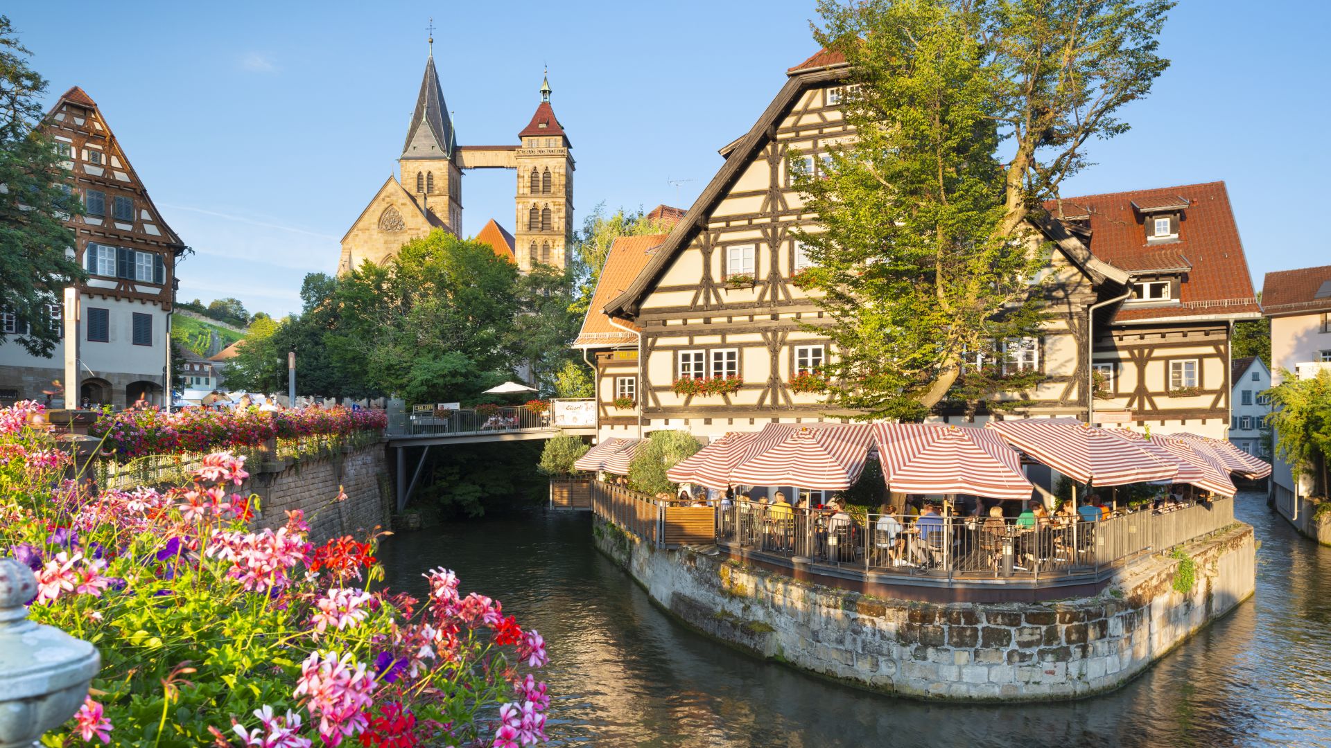 Une maison à colombages pittoresque avec une terrasse de restaurant au bord de l`eau, des fleurs multicolores au premier plan et l`église municipale St Dionys en arrière-plan
