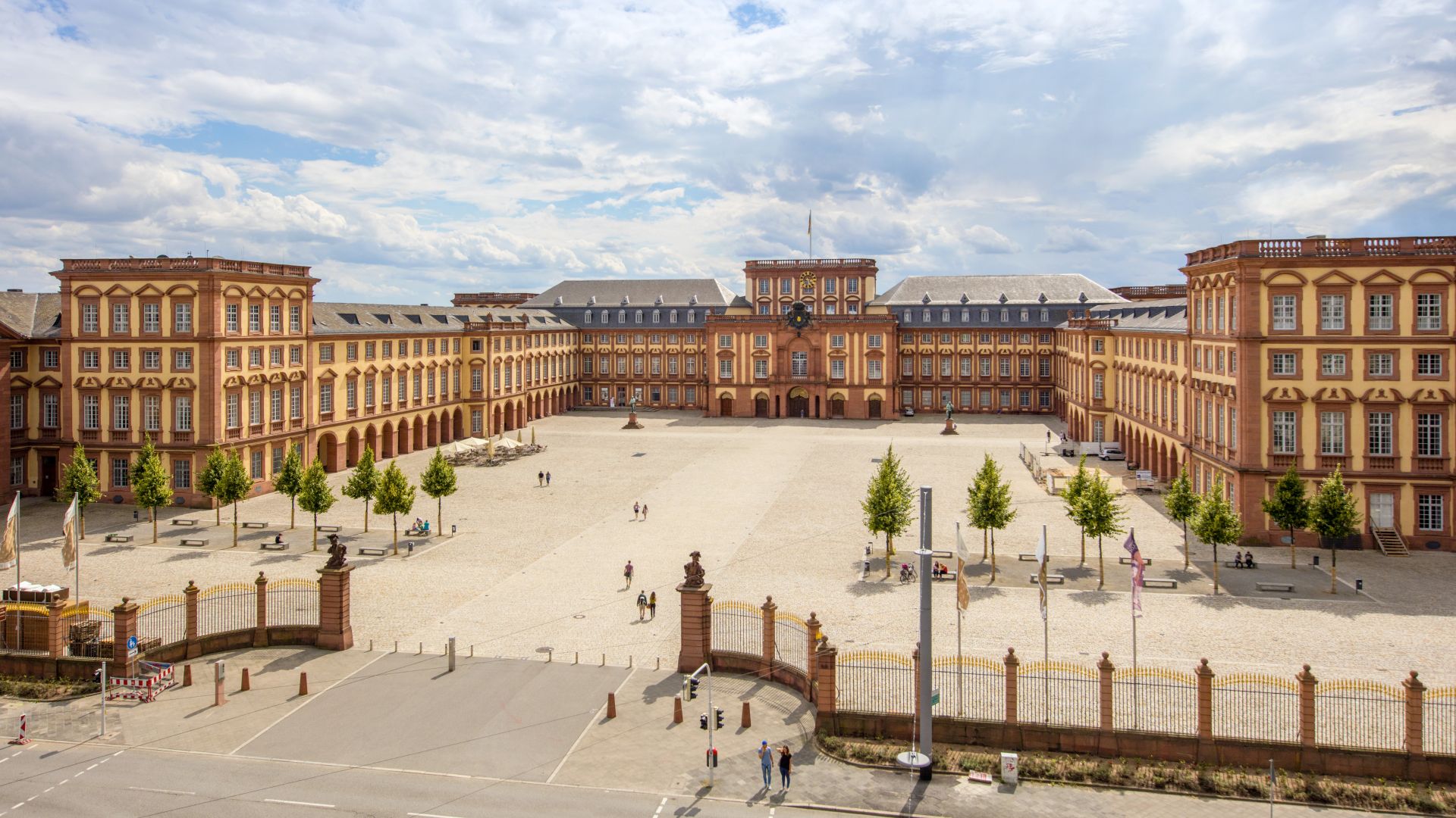 Das Mannheimer Barockschloss steht in U-Form unter einem teilweise bewölkten Himmel. Das Gebäude besteht aus rotem und cremefarbenem Mauerwerk.