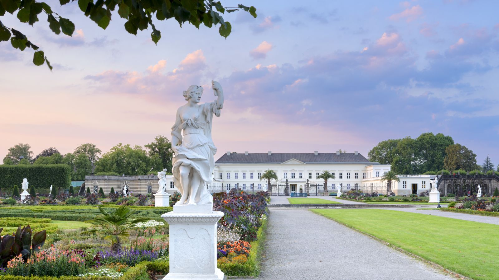 Jardin baroque des Jardins de Herrenhausen, avec une statue au premier plan, des parterres de fleurs et le château au bout de l'allée centrale, sous le ciel du soir