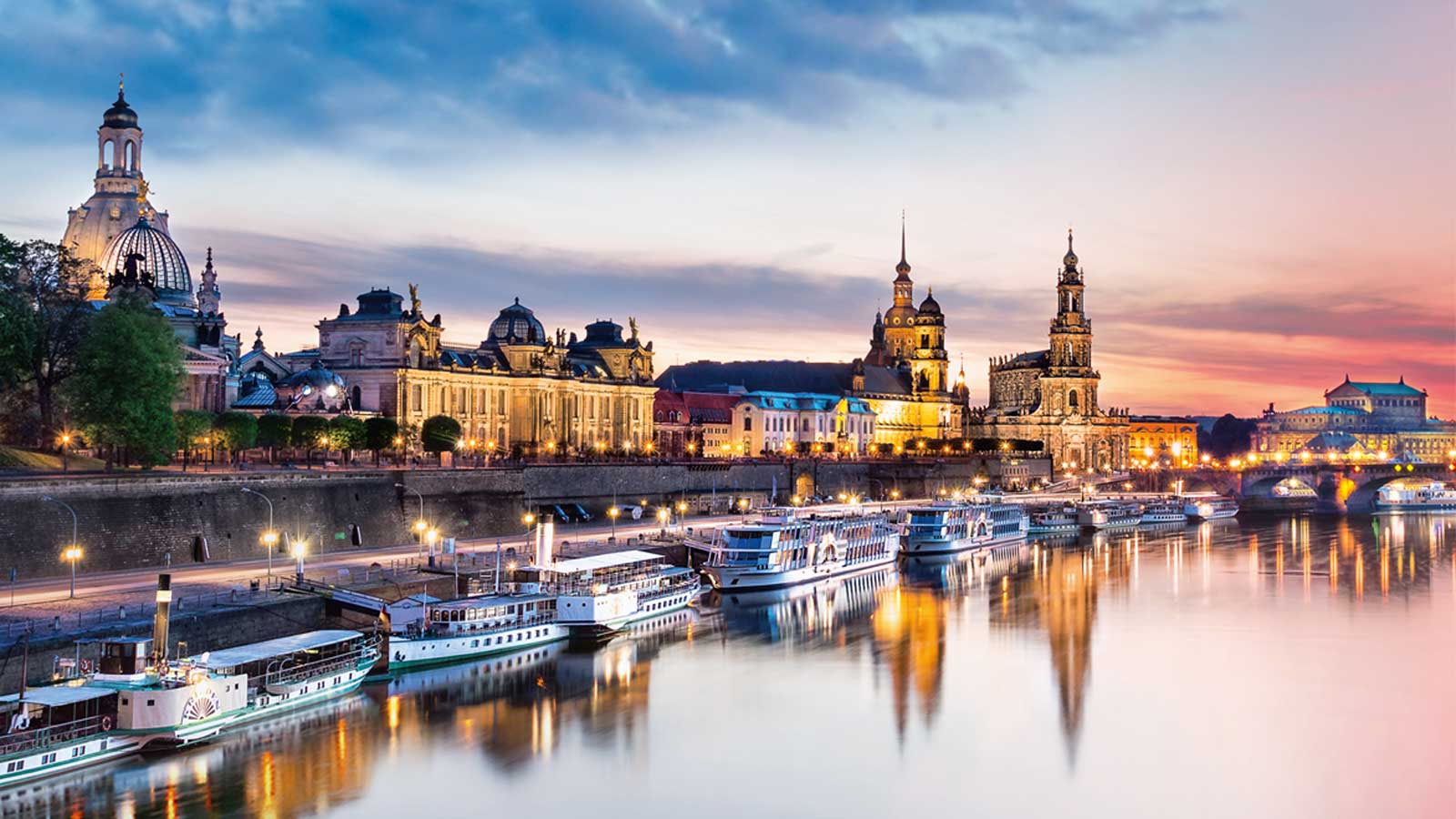 Le soir, plusieurs bateaux sont amarrés sur les rives de l'Elbe à Dresde. Des bâtiments dotés de coupoles et de tours bordent les rives. Le ciel présente un mélange de nuances de bleu et de rose.