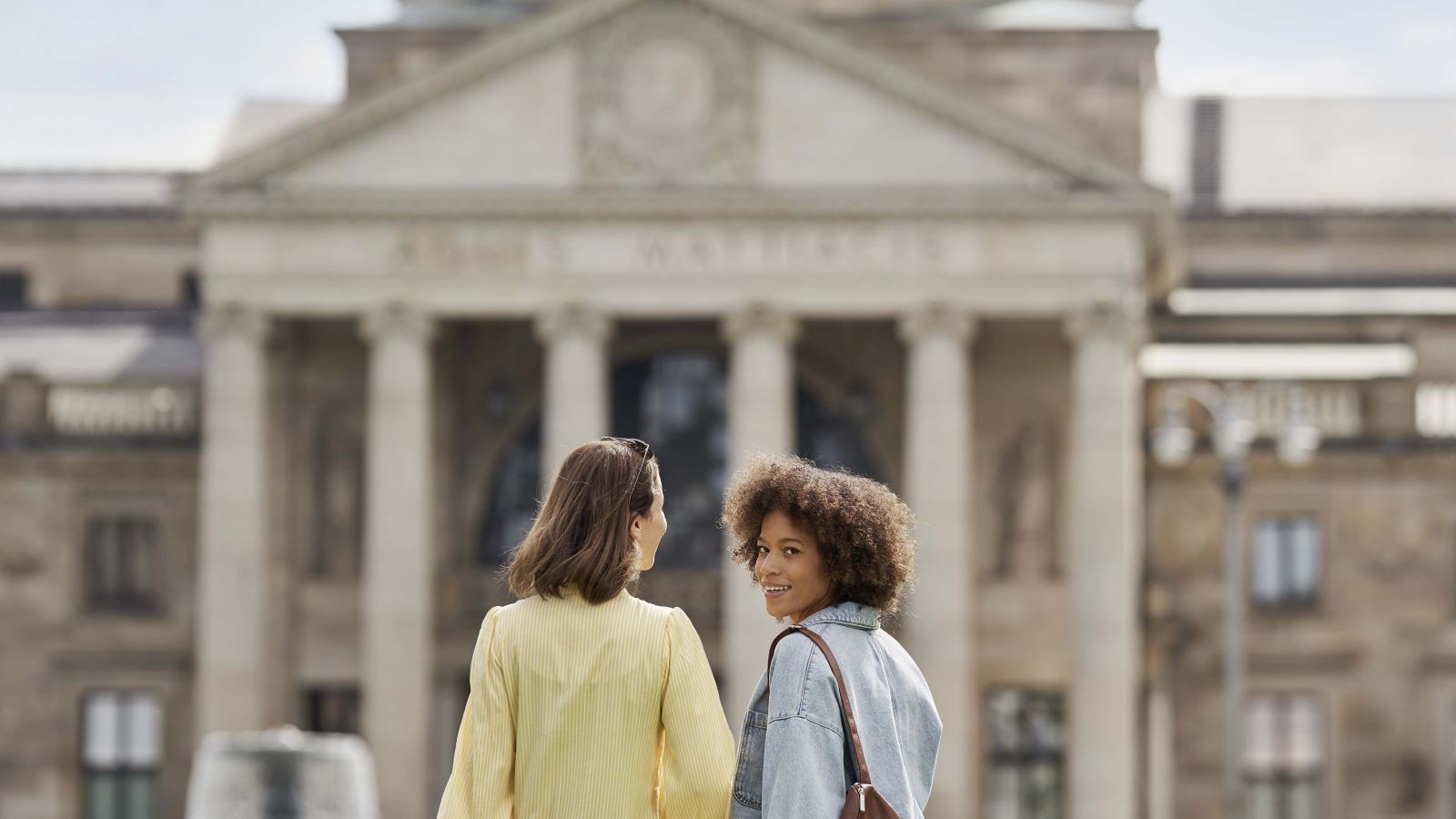Deux femmes se promènent devant le Kurhaus à Wiesbaden. On aperçoit une fontaine à l'arrière-plan.