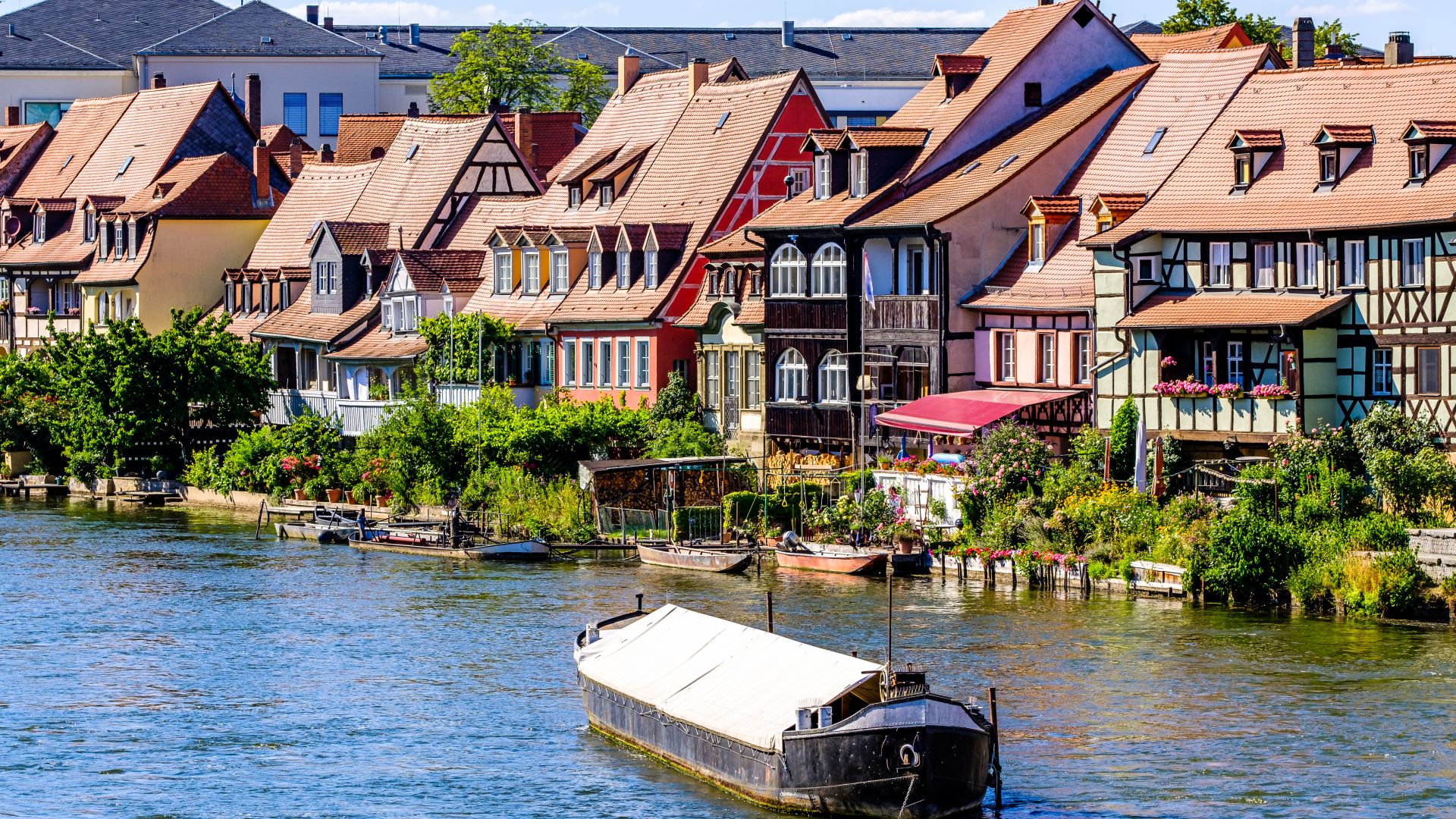 In Bamberg reihen sich entlang der Regnitz bunte Fachwerkhäuser aneinander. Ein Boot mit weißer Plane liegt im Fluss.