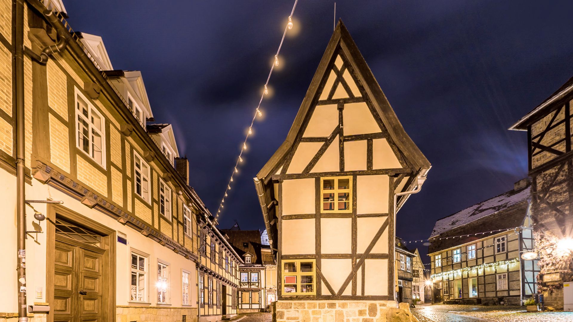 Nachtaufnahme im Winter in Quedlinburg. Es zeigt traditionelle Fachwerkhäuser, die von Straßenlaternen und Lichterketten beleuchtet werden. Im Vordergrund ist die kopfsteingepflasterte Straße zu sehen.