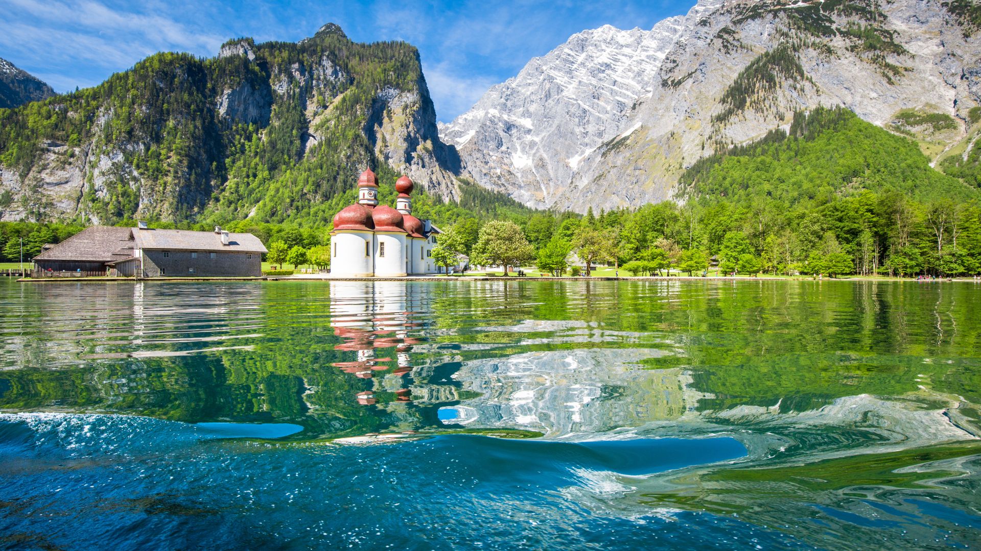 In Schönau steht die Wallfahrtskirche St. Bartholomä am Rande des Königssees. Die Kirche hat rote, zwiebelförmige Kuppeln. Im klaren Wasser spiegeln sich die Kirche, üppig grüne Bäume und die felsigen Berge im Hintergrund.