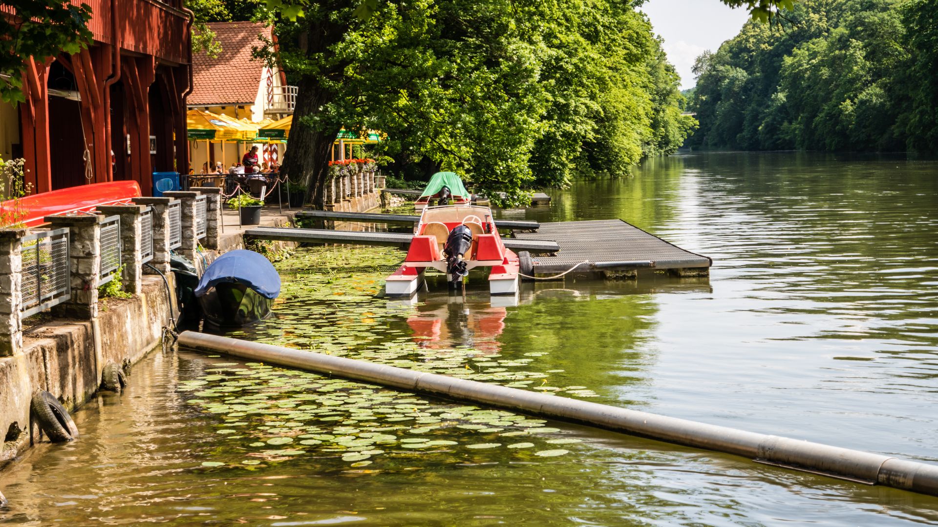 View of an idyllic bathing spot by the river in Hainpark Bamberg with jetties, pedal boat, water lilies and shady trees. In the background, people sit under yellow parasols in front of a café.