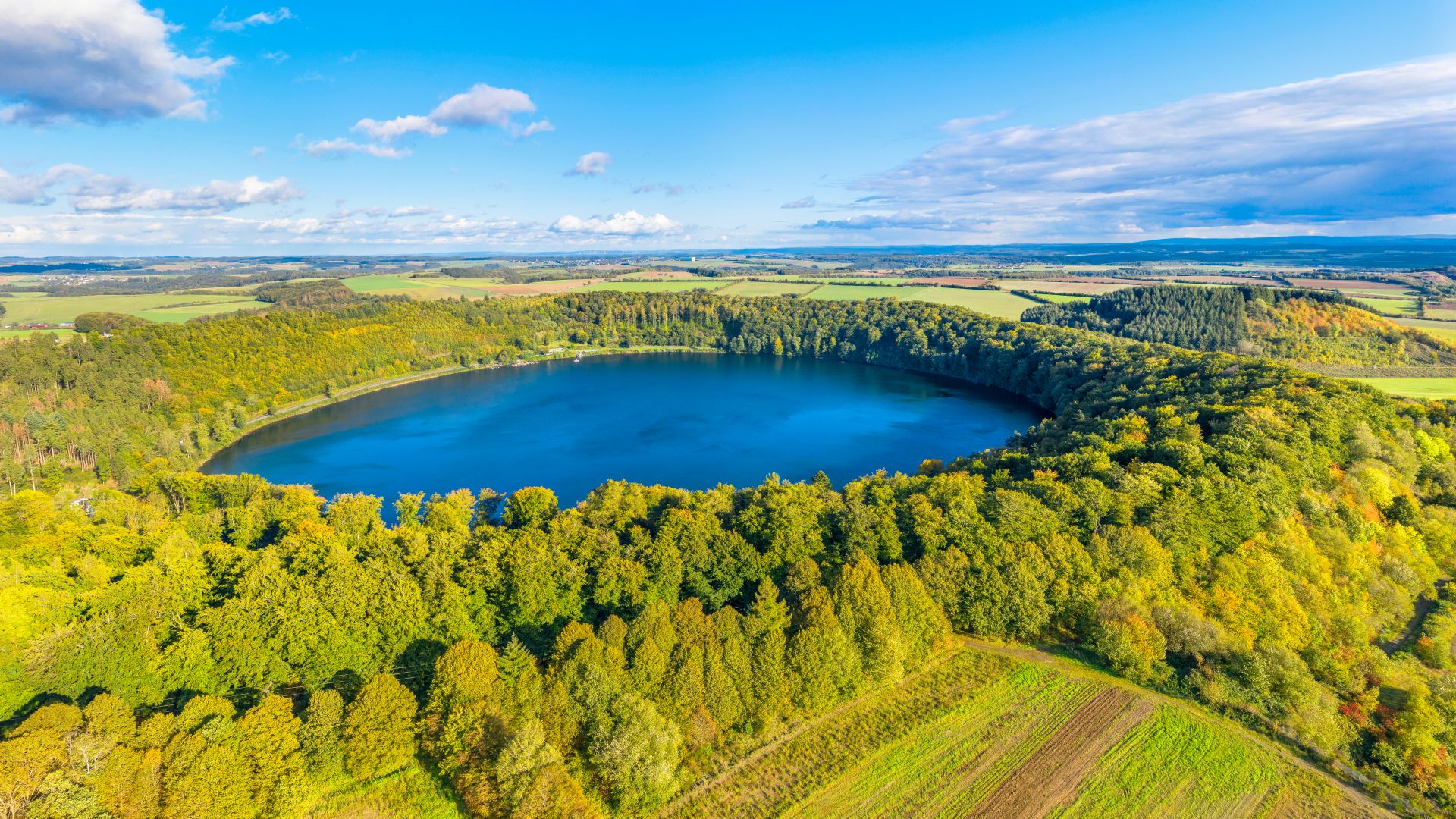 Bird`s eye view of the Pulvermaar crater lake, surrounded by autumn-colored forest and wide fields under a blue sky with loose clouds