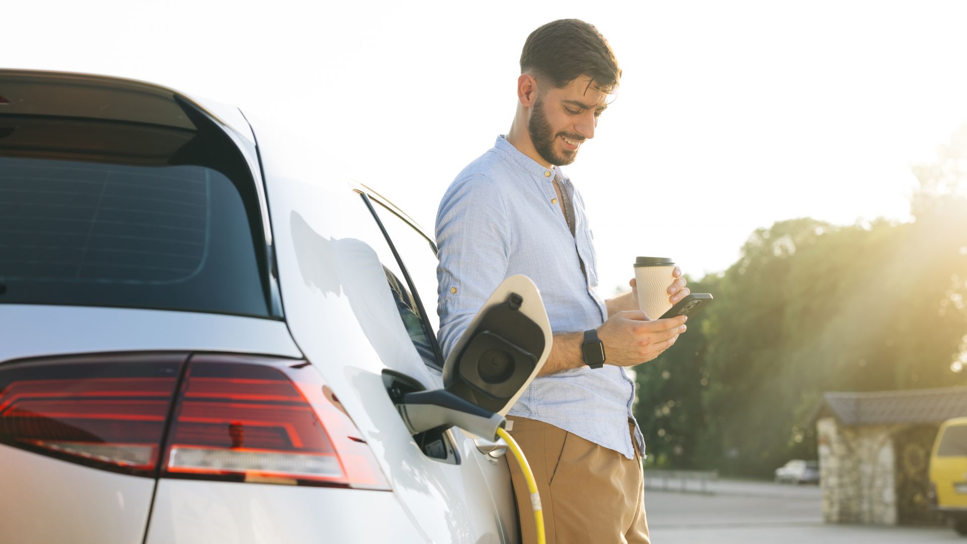 Mann mit Smartphone und Kaffee steht mit Elektroauto an Ladesäule