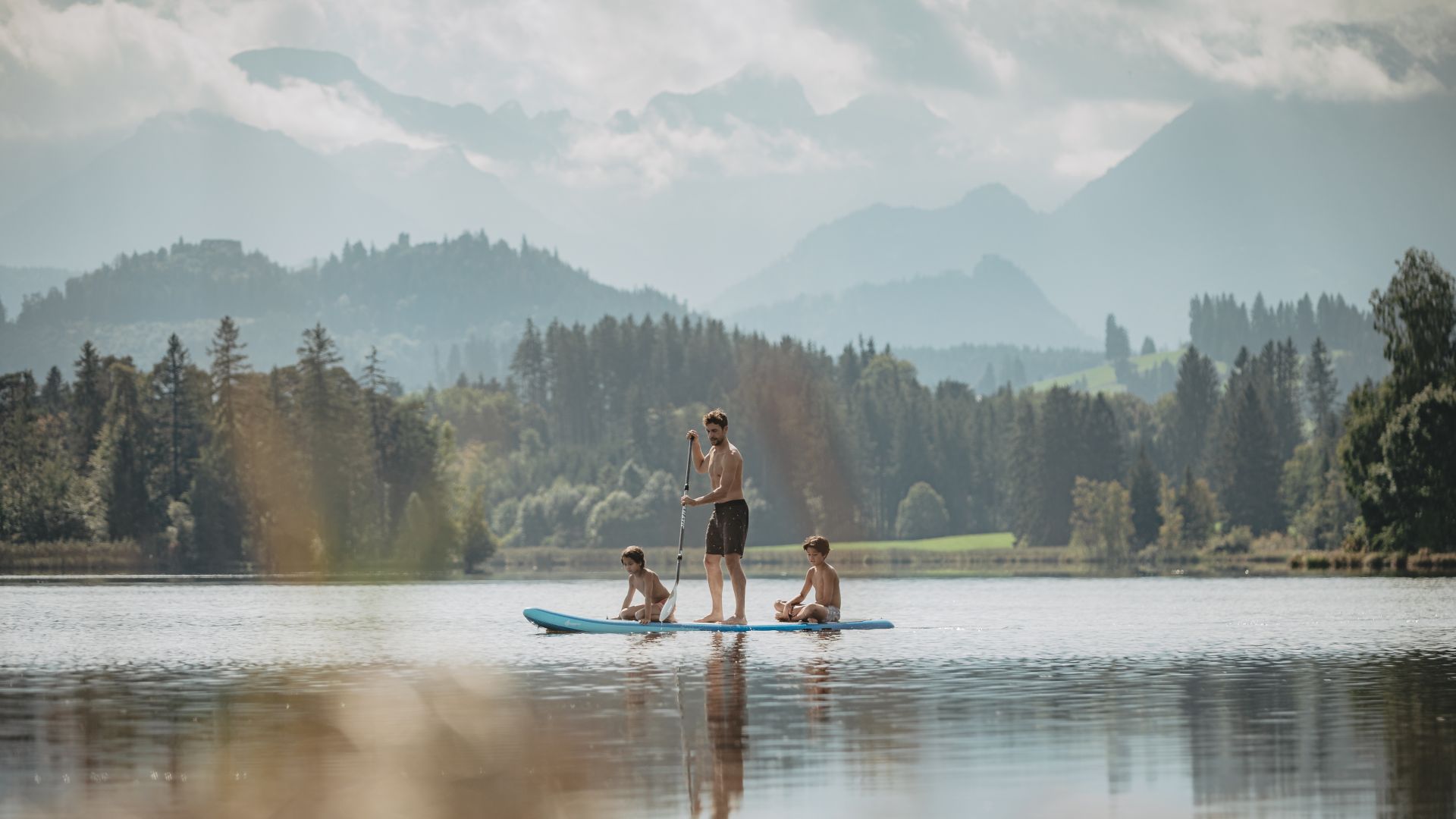 Nesselwang : Famille s'amusant avec une planche de surf au Schwaltenweiher dans les Préalpes avec les Alpes d'Allgäu en arrière-plan