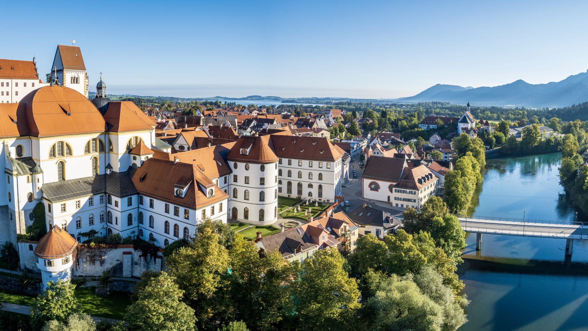Gebäude mit roten Dächern in der Stadt Füssen stehen inmitten von grünen Bäumen links neben dem Fluss Lech.