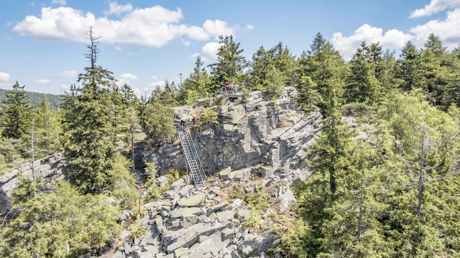 Felsenlandschaft mit einem Aussichtspunkt aus Metall, umgeben von dichten Nadelbäumen, unter blauem Himmel mit vereinzelten Wolken. Eine Metalltreppe führt zum Aussichtspunkt, auf dem mehrere Personen stehen. Die Szene spielt sich in einer bewaldeten Berglandschaft ab.
