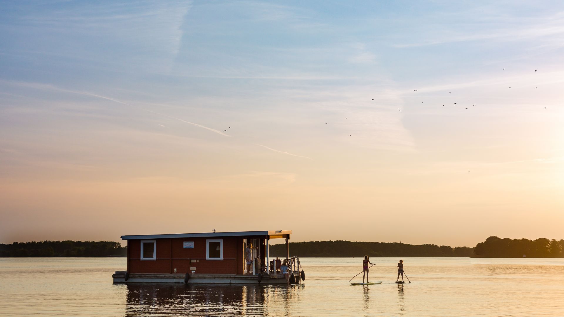 Ein Hausboot schwimmt bei Sonnenuntergang auf der Müritz in der Region Mecklenburgische Seenplatte. In der Nähe des Hausbootes sind zwei Personen auf einem Stand-up-Paddleboard unterwegs.