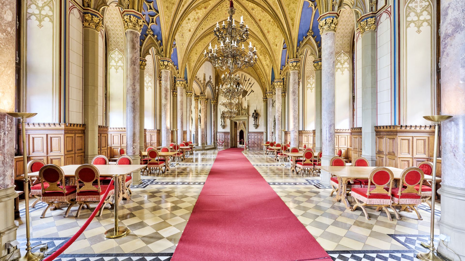 Grafensaal in der Burg Hohenzollern in Hechingen. Der Saal hat hohe, gewölbte Decken, die mit goldenen Mustern und blauen Tafeln verziert sind. An den Seiten des Saals befinden sich große Marmorsäulen. Der Wandelgang ist mit roten Teppichen ausgelegt, und von der Decke hängen kunstvolle Kronleuchter. Die unteren Wände sind holzgetäfelt, und an den Seiten des Saals sind Tische mit roten Stühlen aufgestellt.