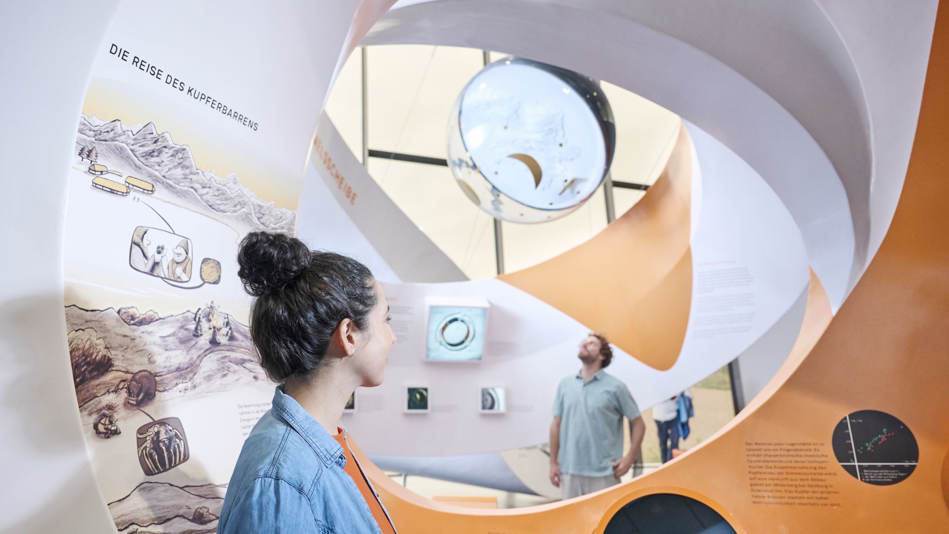 Une femme regarde l`exposition sur le voyage du lingot de cuivre, tandis qu`un homme admire la maquette du disque céleste à l`arrière-plan.