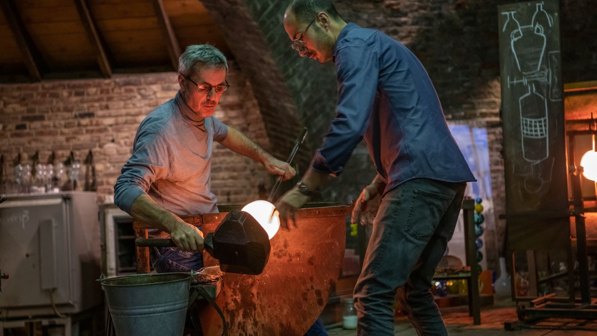Zwei Männer arbeiten in einer Glasbläserei. Einer hält eine lange Pfeife mit glühendem Glas, während der andere assistiert. Sie befinden sich in einer rustikalen, ziegelverklinkerten Werkstatt mit hölzerner Decke. Rechts ist eine Tafel mit Skizzen.