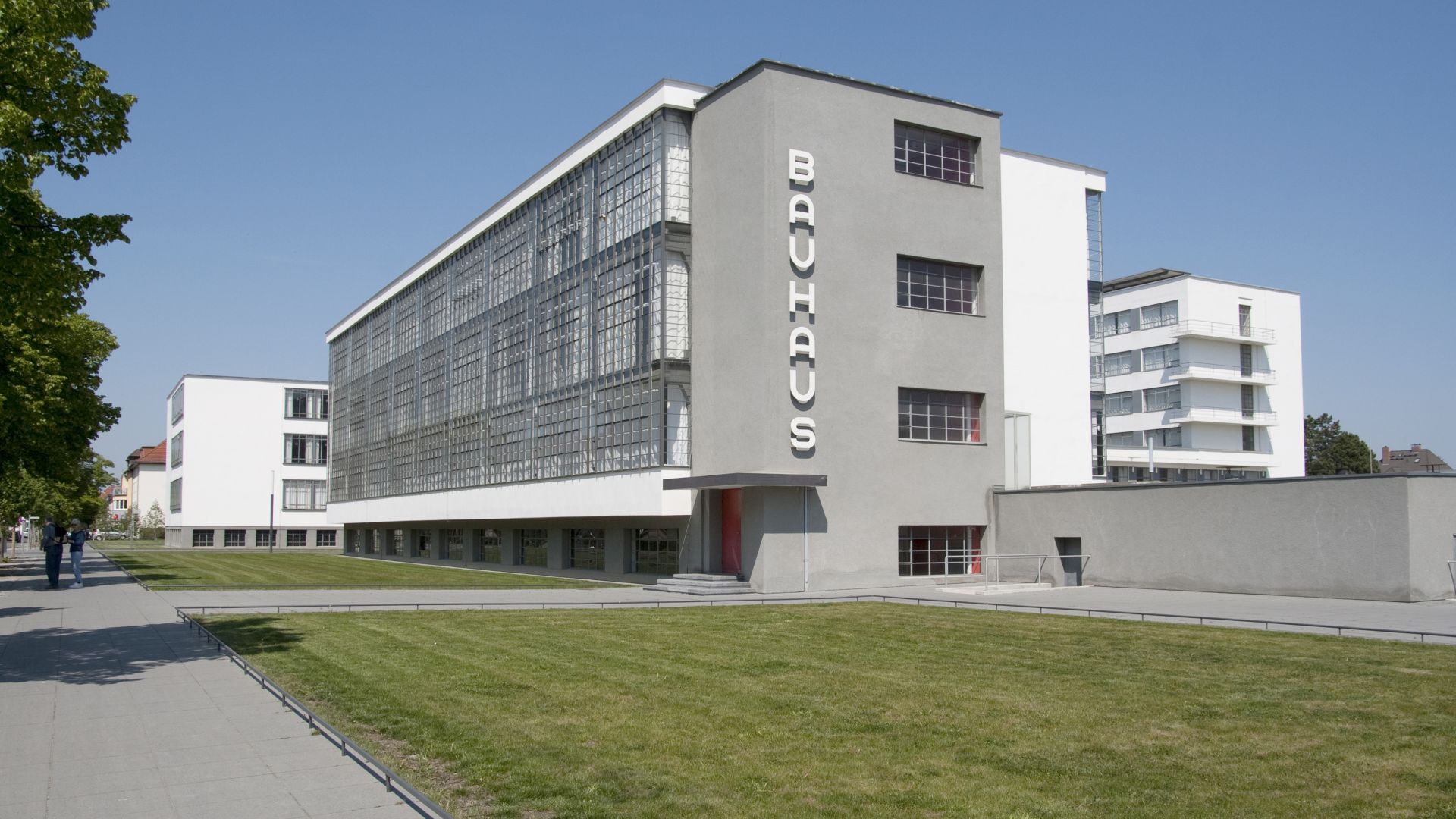 Ein modernes Gebäude in Dessau-Roßlau, das 1925/26 vom Architekten Walter Gropius entworfen wurde. Das Gebäude ist Teil des Bauhauses, das zum UNESCO-Weltkulturerbe gehört. Es ist bekannt für sein weiß-graues Äußeres mit großen Fenstern und einem Flachdach. Im Vordergrund verläuft ein gepflasterter Gehweg entlang einer grünen Rasenfläche.