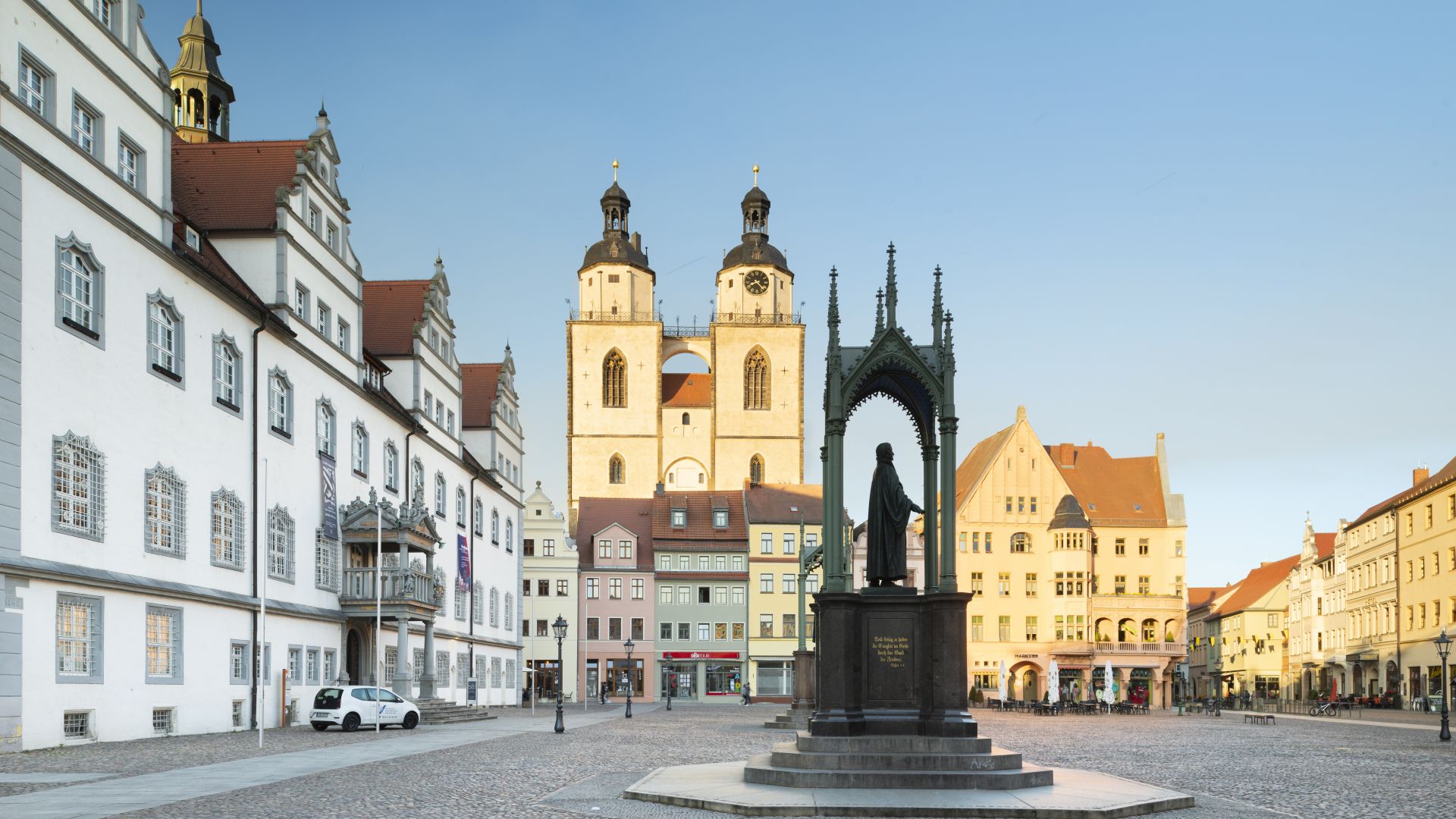 Der Marktplatz in Wittenberg ist mit Kopfsteinpflaster gepflastert. Um den Platz herum befinden sich historische Gebäude und Geschäfte. Im Hintergrund steht die Stadtkirche St. Marien mit zwei markanten Türmen. Im Vordergrund befindet sich ein Denkmal mit einer Statue. Die Gebäude haben einen ähnlichen Baustil mit verzierten Fenstern und roten Dächern. Die Gebäude rechts und im Hintergrund werden von der Sonne angestrahlt, die Fassade links liegt im Schatten. Der Himmel ist blau und wolkenlos.