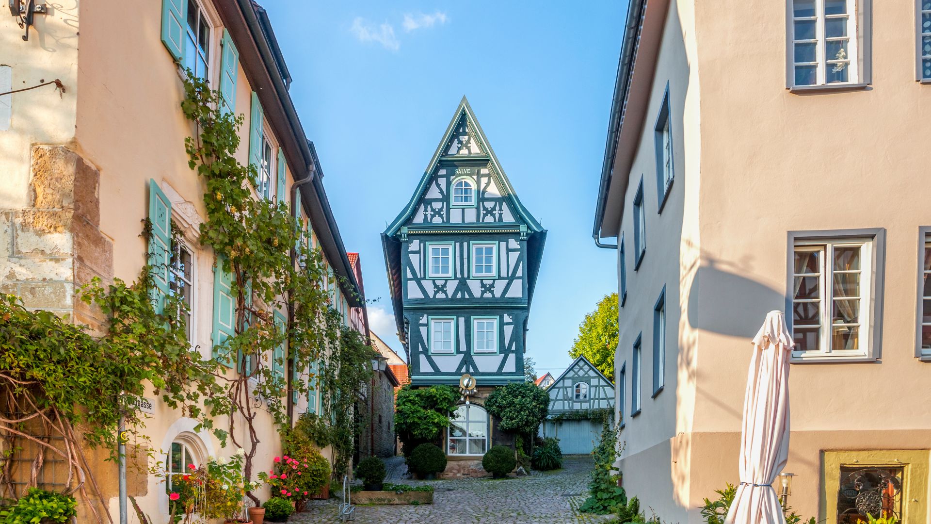 Eine kopfsteingepflasterte Gasse in Bad Wimpfen wird von zwei Gebäuden mit bunten Fensterläden und Kletterpflanzen flankiert. Am Ende der Gasse steht ein hohes, schmales Fachwerkhaus mit steilem, spitzem Dach und kunstvollen Verzierungen. Im Hintergrund sind Bäume und ein weiteres Fachwerkhaus unter blauem Himmel zu sehen.