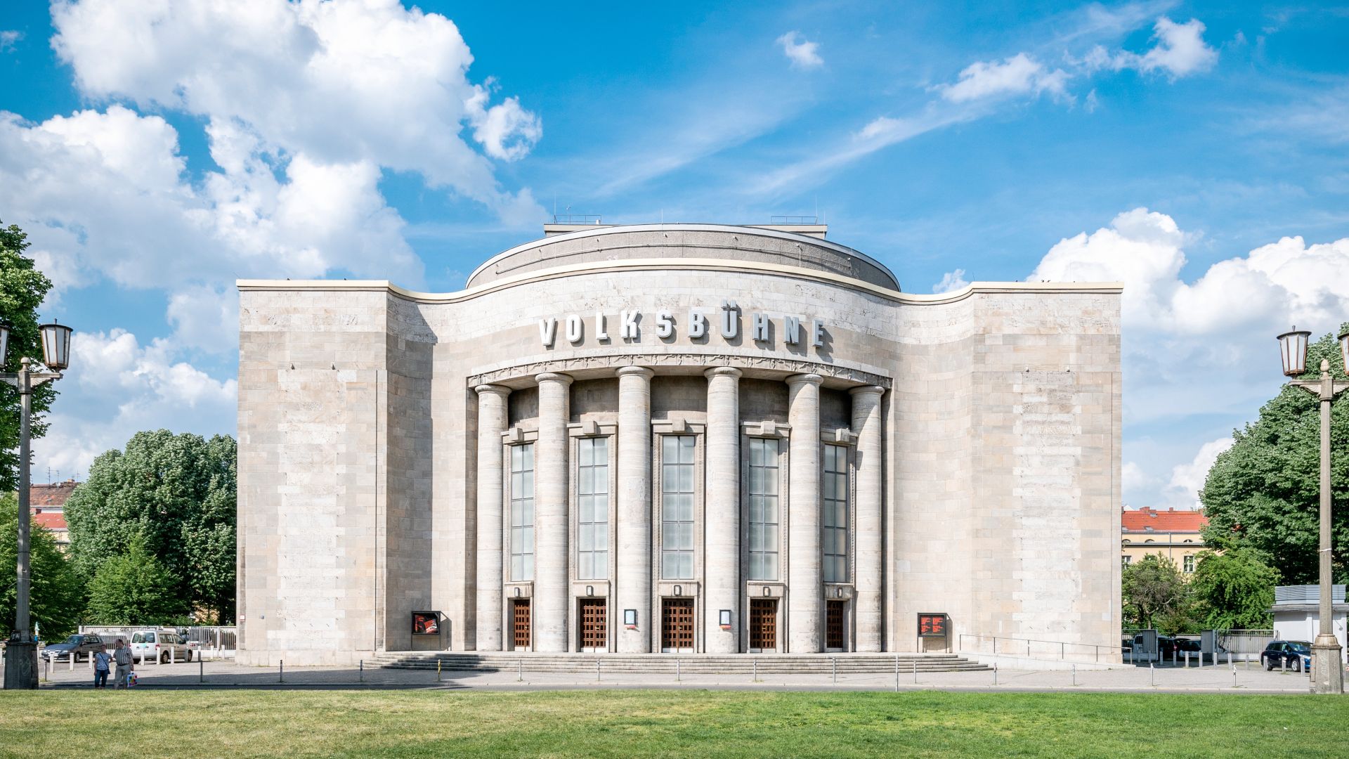 Das Bild zeigt die Volksbühne, ein markantes Gebäude in einer Stadt. Der Bau ist aus hellem Stein mit großen Säulen und dem Schriftzug 