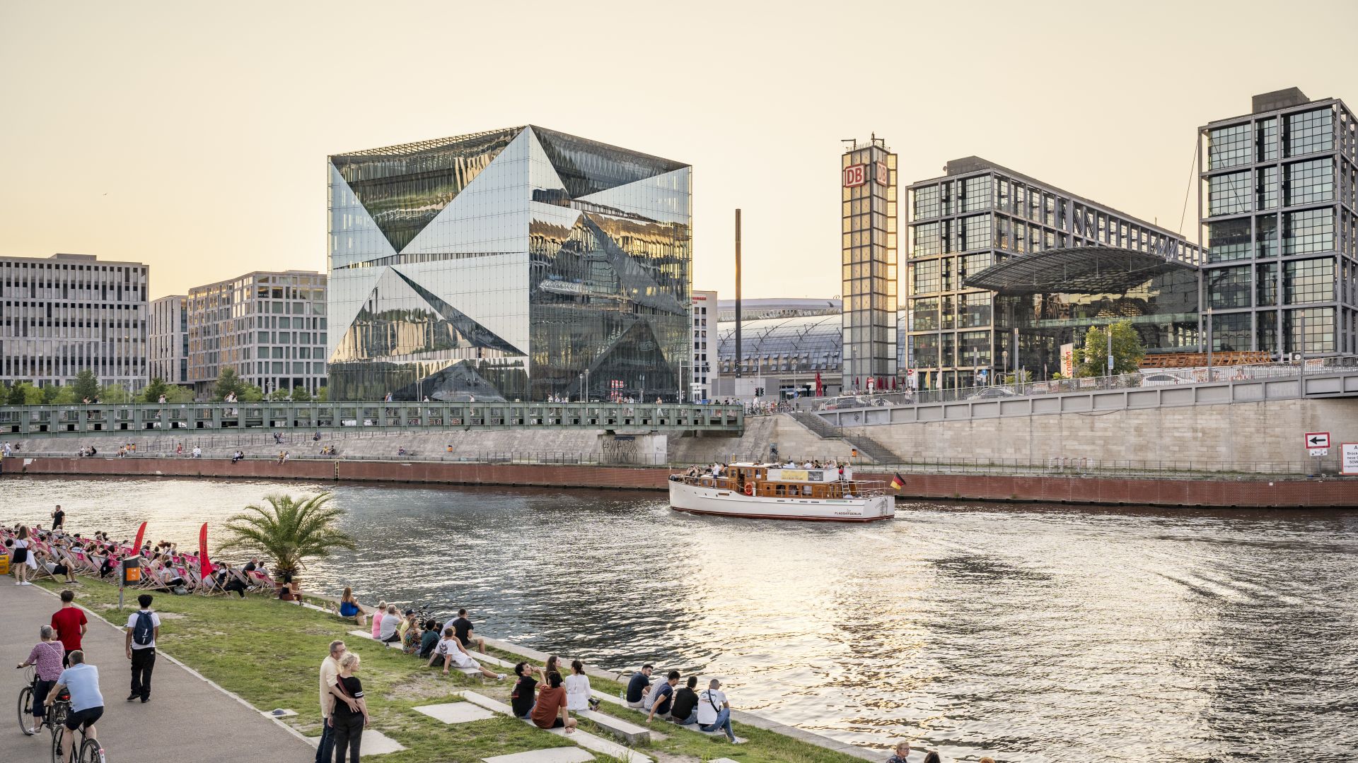 Des personnes se promènent sur les rives de la Spree avec le Cube Berlin et la gare centrale de Berlin en arrière-plan, tandis qu`un bateau passe.