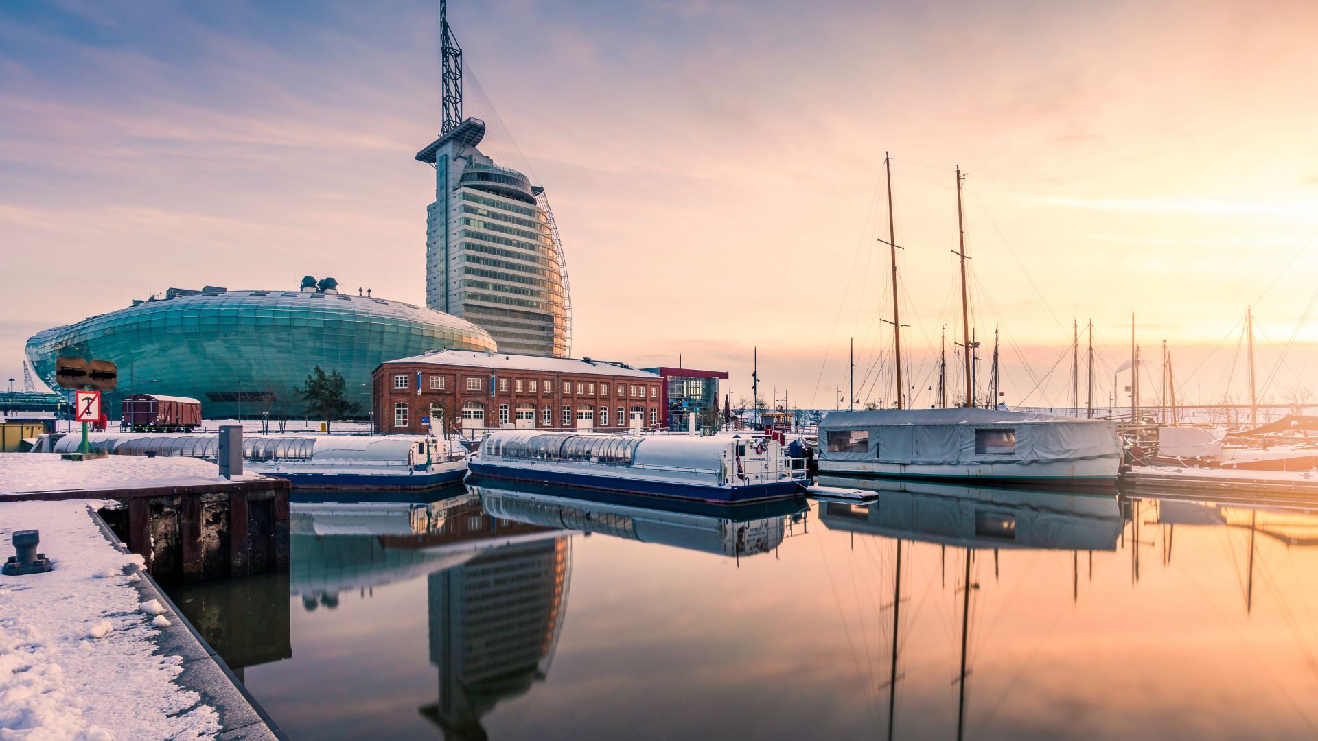 Neuer Hafen in Bremerhaven im Winter mit dem Klimahaus und Segelbooten. Die Uferpromenade ist schneebedeckt, und im Wasser spiegeln sich die Gebäude und Boote. Der Himmel zeigt die Pastellfarben des Sonnenaufgangs oder Sonnenuntergangs.