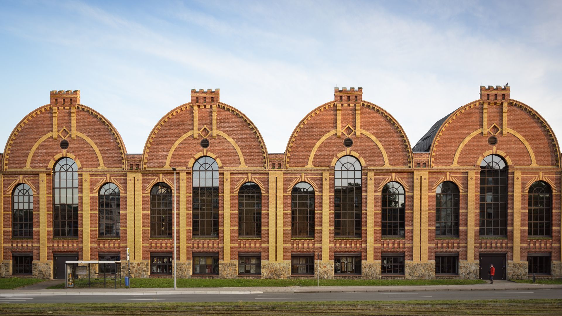 Sächsisches Industriemuseum in Chemnitz verfügt über ein großes Backsteingebäude mit vier identischen Bogenteilen. Vor dem Gebäude befinden sich eine Wiese und eine Straße mit Straßenbahnschienen.