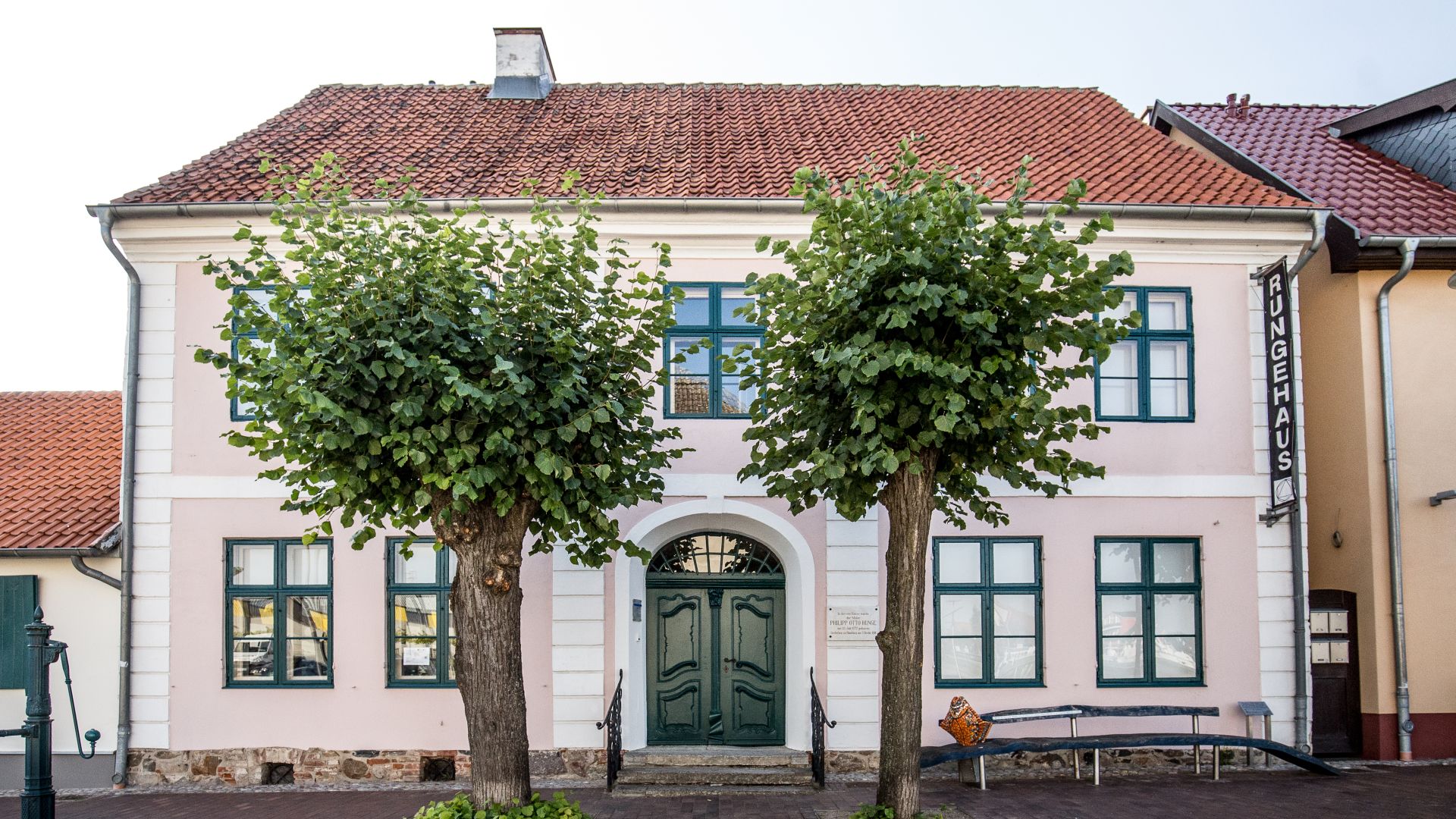 Front view of the two-story Runge House in Wolgast, with a light-colored exterior, green-framed windows, and two trees in front of the entrance.