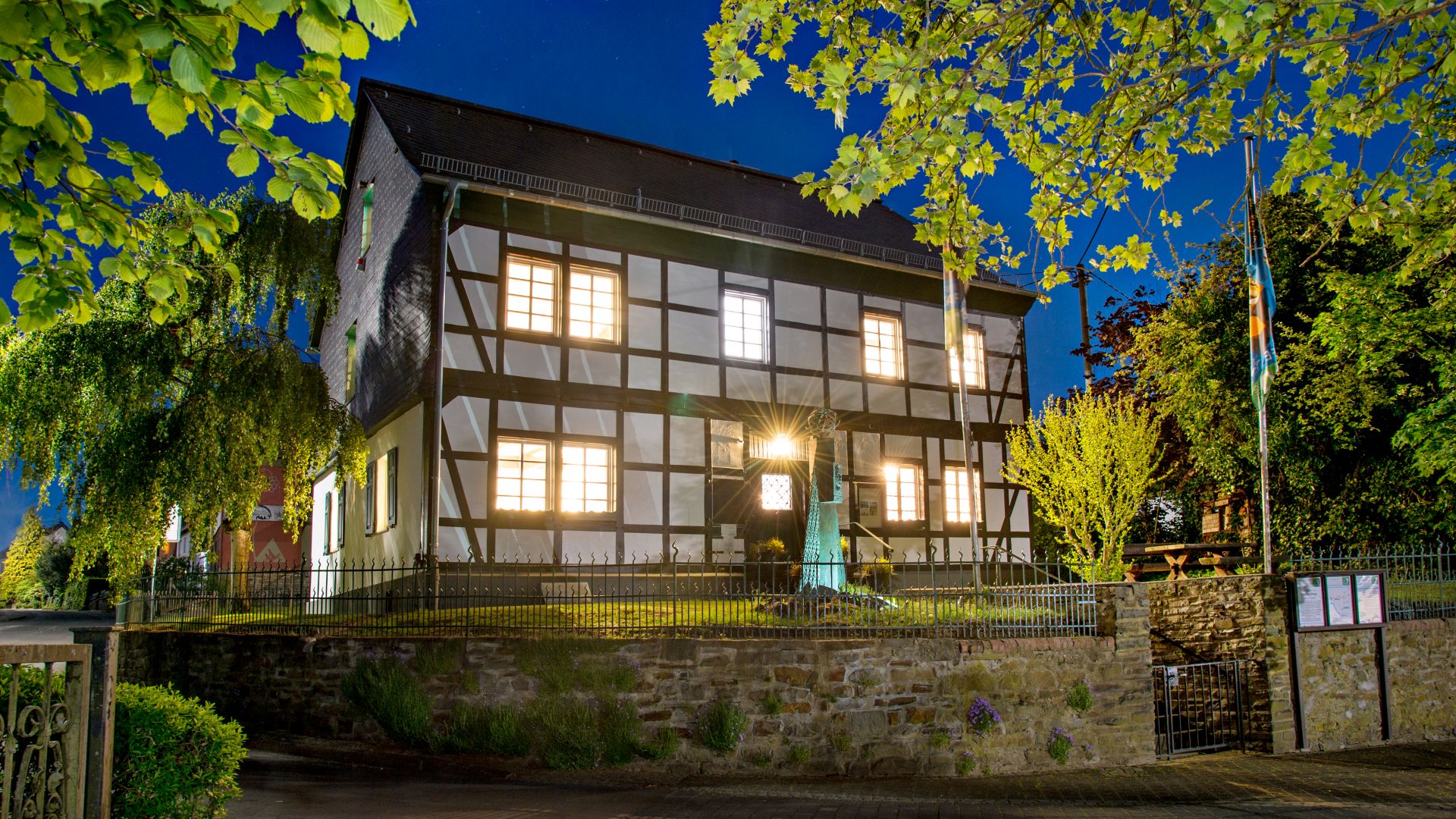 Ein beleuchtetes Fachwerkhaus bei Nacht mit einem kleinen Garten, umgeben von Bäumen, vermutlich in Deutschland. Das Gebäude ist von einem niedrigen Zaun umgeben und liegt an einer gepflasterten Straße.