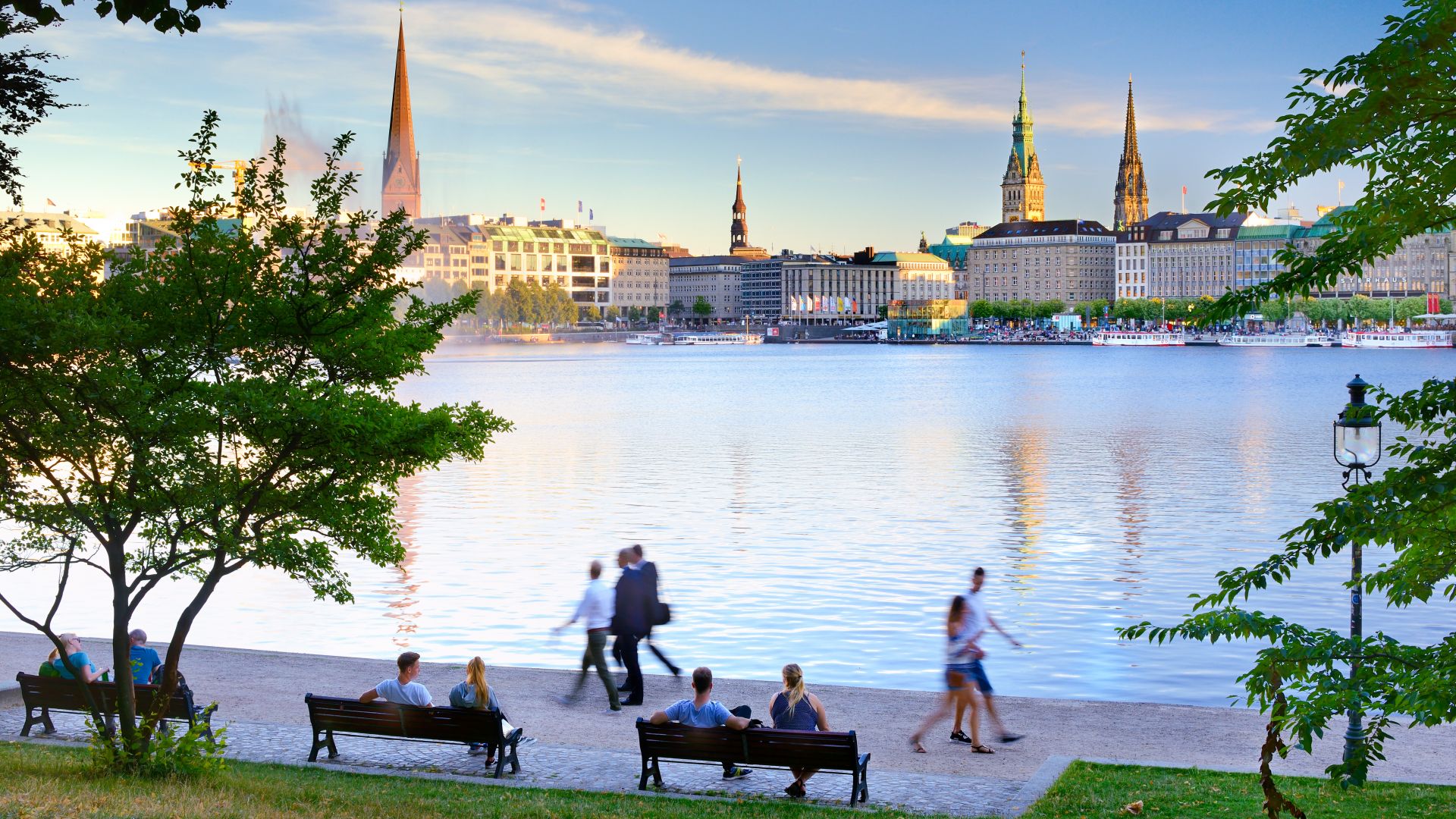 Spaziergänger und Sitzbänke an der Binnenalster mit Blick auf Hamburgs Altstadtkulisse im Abendlicht