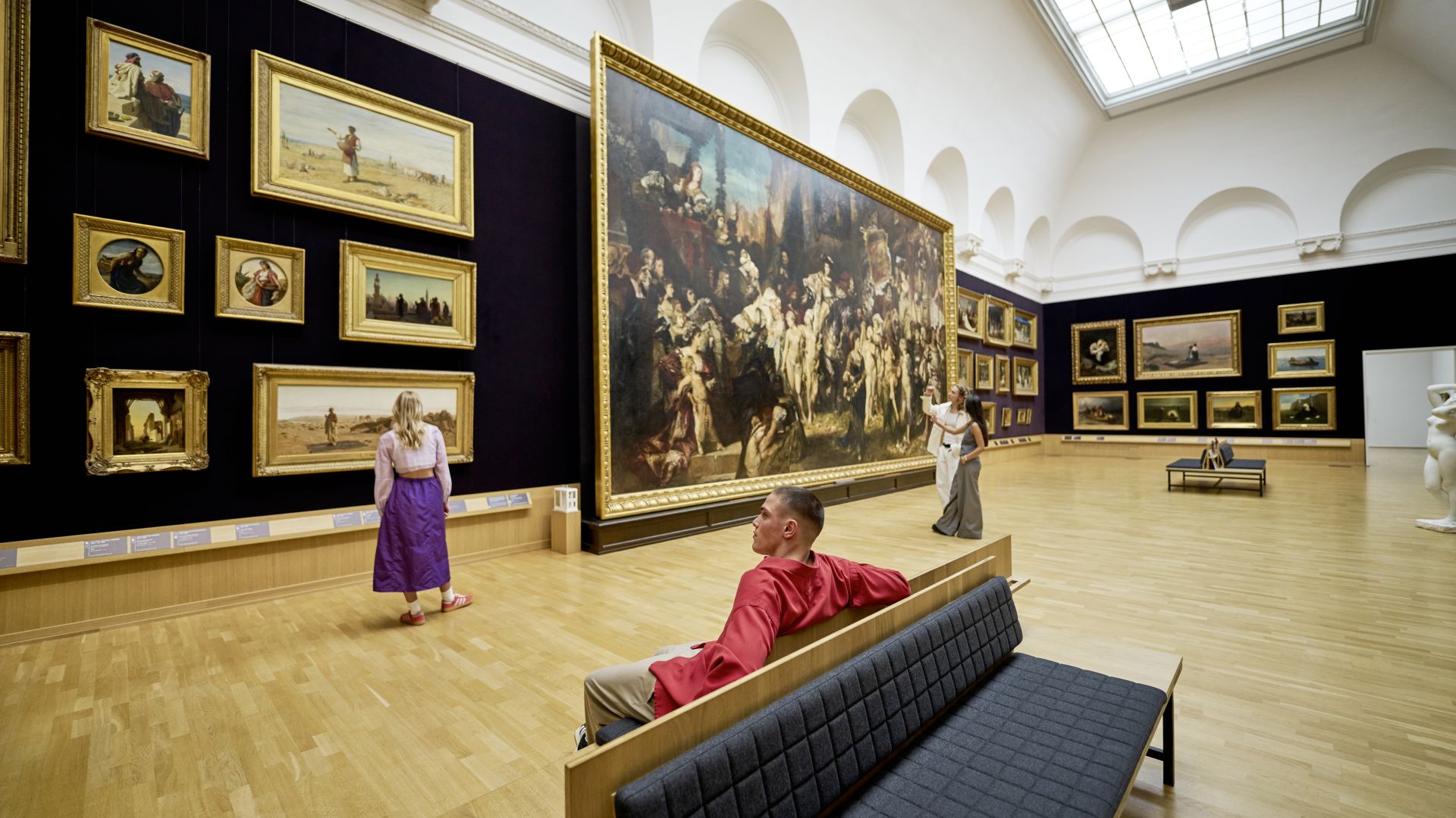 In einem lichtdurchfluteten Museumsraum betrachten Personen Gemälde, die an einer Wand in vergoldeten Rahmen hängen. Ein großes, zentrales Gemälde zieht besondere Aufmerksamkeit auf sich. Einige Besucher:innen sitzen auf Bänken, während andere die Kunstwerke aus nächster Nähe betrachten.