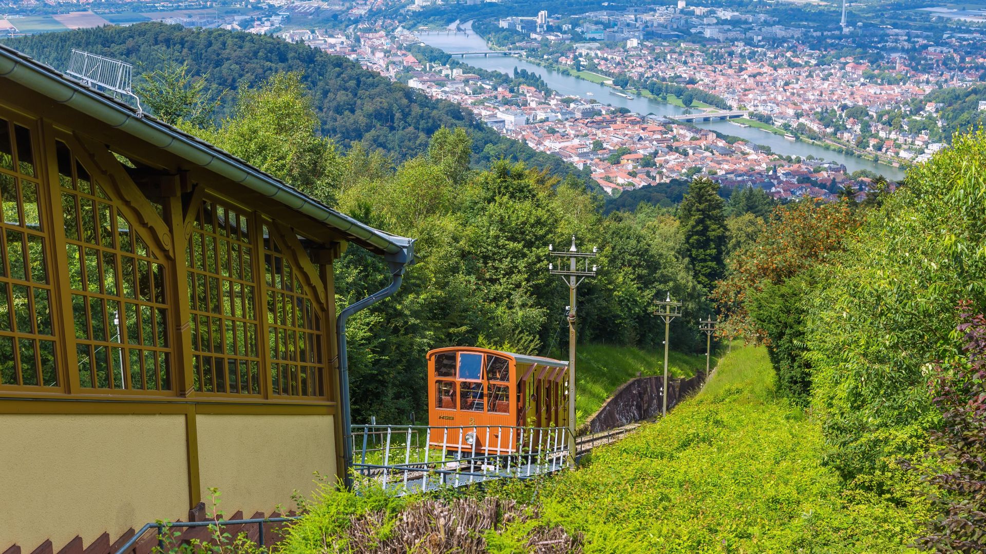 Ein orangefarbener Waggon fährt in der Nähe einer Bergstation der Königstuhlbahn und Standseilbahn, mit einer schönen Aussicht auf die Stadt Heidelberg.