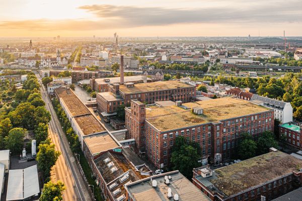 Ein Luftbild von Leipzig während der Dämmerung zeigt den Komplex der Baumwollspinnerei mit seinen großen Backsteingebäuden. Die Straßen und die Umgebung sind begrünt und in der Ferne erstrecken sich weitere Gebäude.