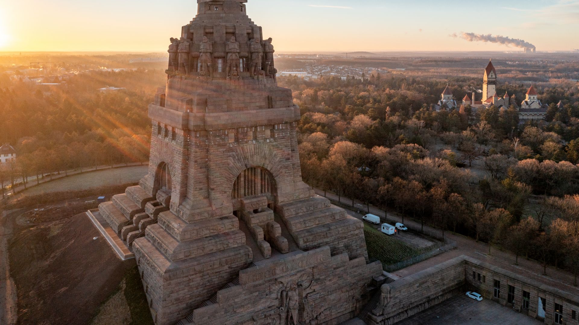 Das Foto zeigt das Völkerschlachtdenkmal in Leipzig aufgenommen in der Abenddämmerung von oben. Ein großes, steinernes Denkmal steht in der Bildmitte, umgeben von Bäumen und einigen Gebäuden. Der Himmel ist klar und die untergehende Sonne wirft ein warmes Licht auf das Denkmal und die Landschaft. Im Hintergrund steigt Rauch aus einem entfernten Industriegebiet auf.
