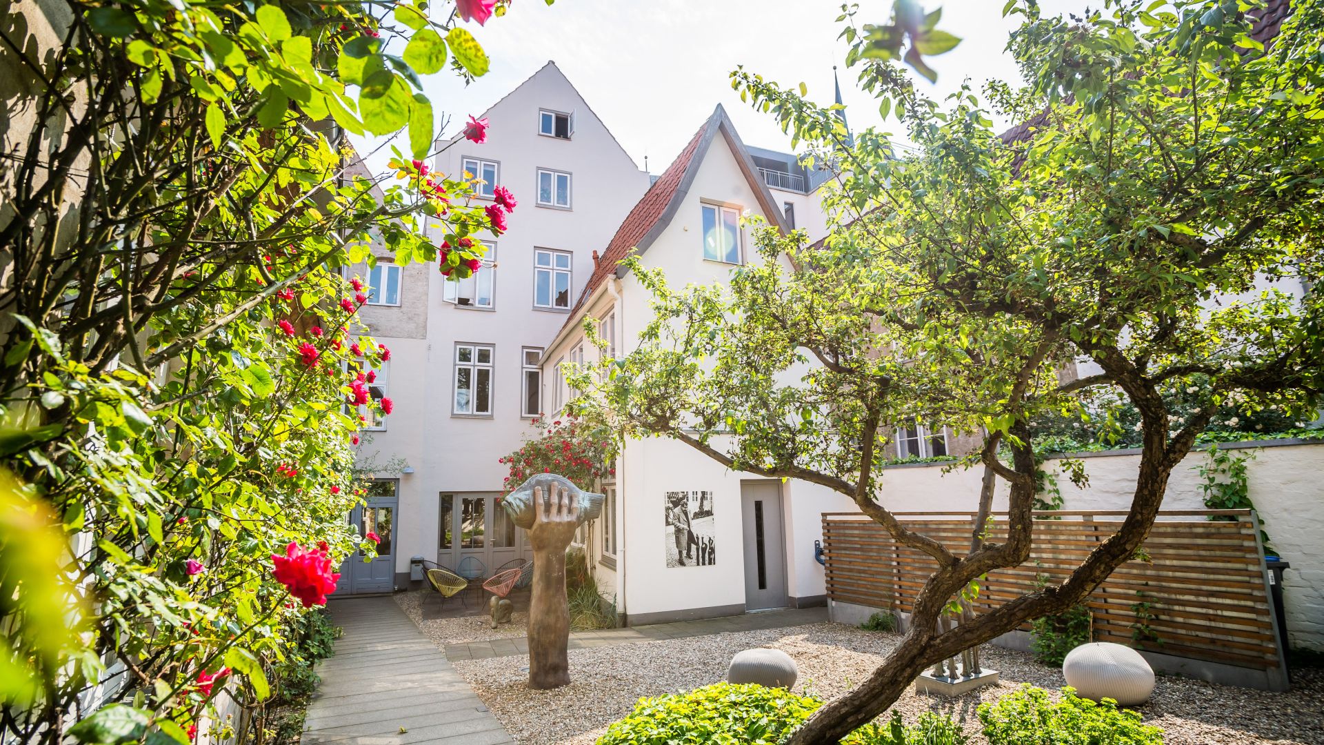Der Innenhof des Günter Grass-Hauses und -Museums in Lübeck ist von einem Weg mit roten Blumen und Sträuchern gesäumt. In der Mitte steht die Skulptur einer großen Hand. Weiße Gebäude mit mehreren Fenstern umgeben den Innenhof. Es gibt Bäume und Sträucher sowie eine Kiesfläche mit dekorativen Steinelementen.