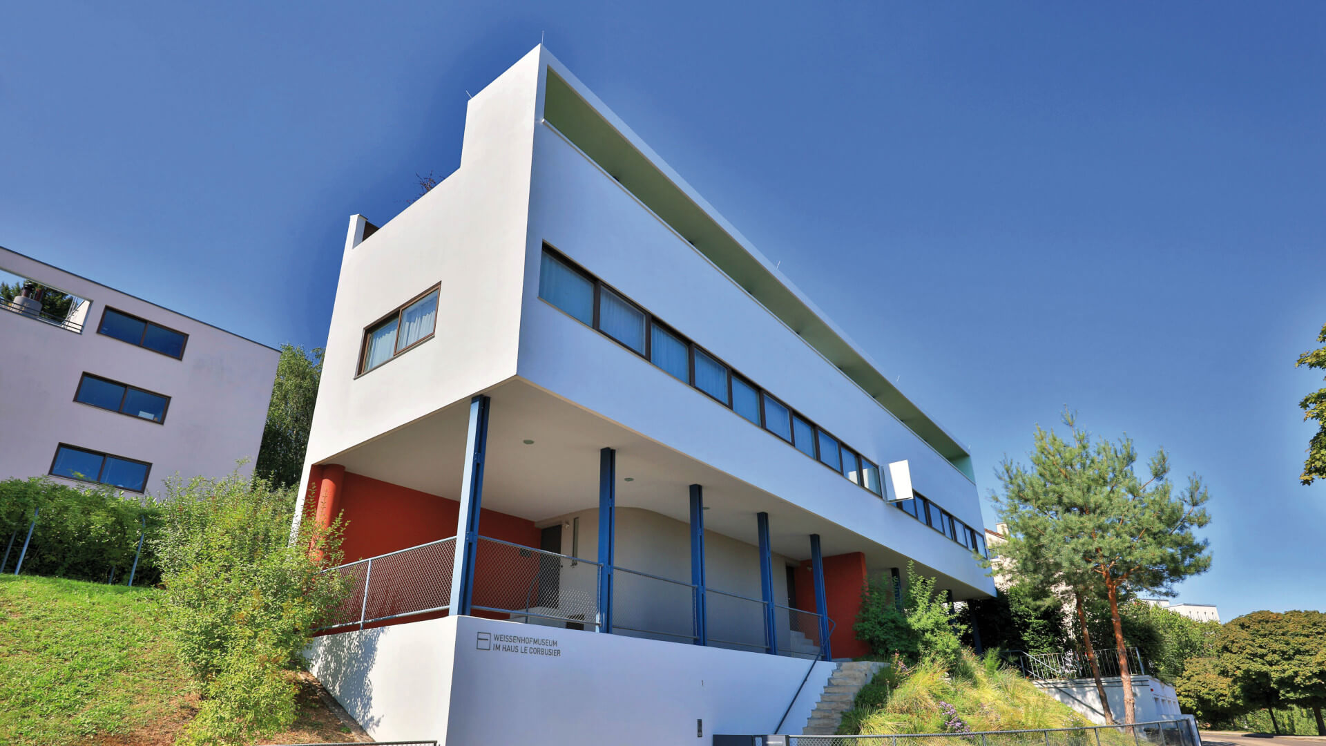 Ein modernes weißes Gebäude mit flachem Dach und klaren Linien ist unter einem blauen Himmel zu sehen. Das 1927 von Le Corbusier und Pierre Jeanneret entworfene Gebäude ist Teil des Landguts und Museums Weissenhof in Stuttgart. Das Gebäude ist begrünt, ein Weg führt zu ihm hinauf, und es sind einige Texteinblendungen mit Copyright-Informationen in Englisch und Deutsch zu sehen.