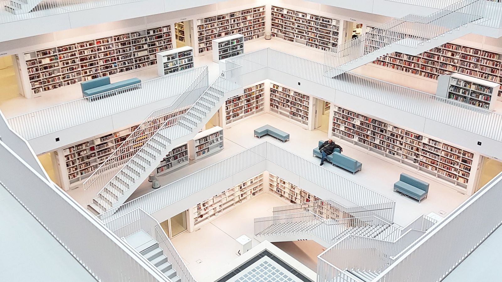 Ein mehrstöckiger Innenraum mit Regalreihen an den Wänden in der Stuttgarter Stadtbibliothek. Die Gestaltung umfasst weiße Geländer, Freitreppen und Sitzbereiche mit Menschen auf verschiedenen Ebenen.