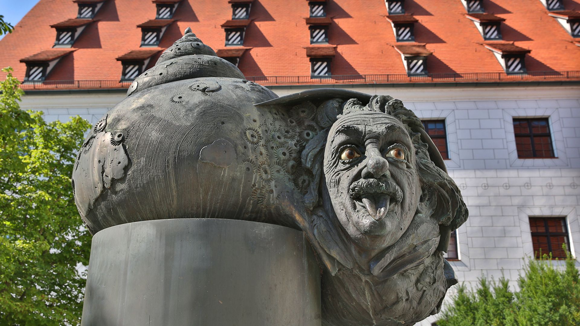 Une sculpture en bronze montre la tête d`Albert Einstein tirant la langue, montée sur un socle lisse et sombre. En arrière-plan, on peut voir un bâtiment aux murs blancs et au toit rouge avec de nombreuses lucarnes. Quelques arbres flanquent la zone autour de la sculpture.