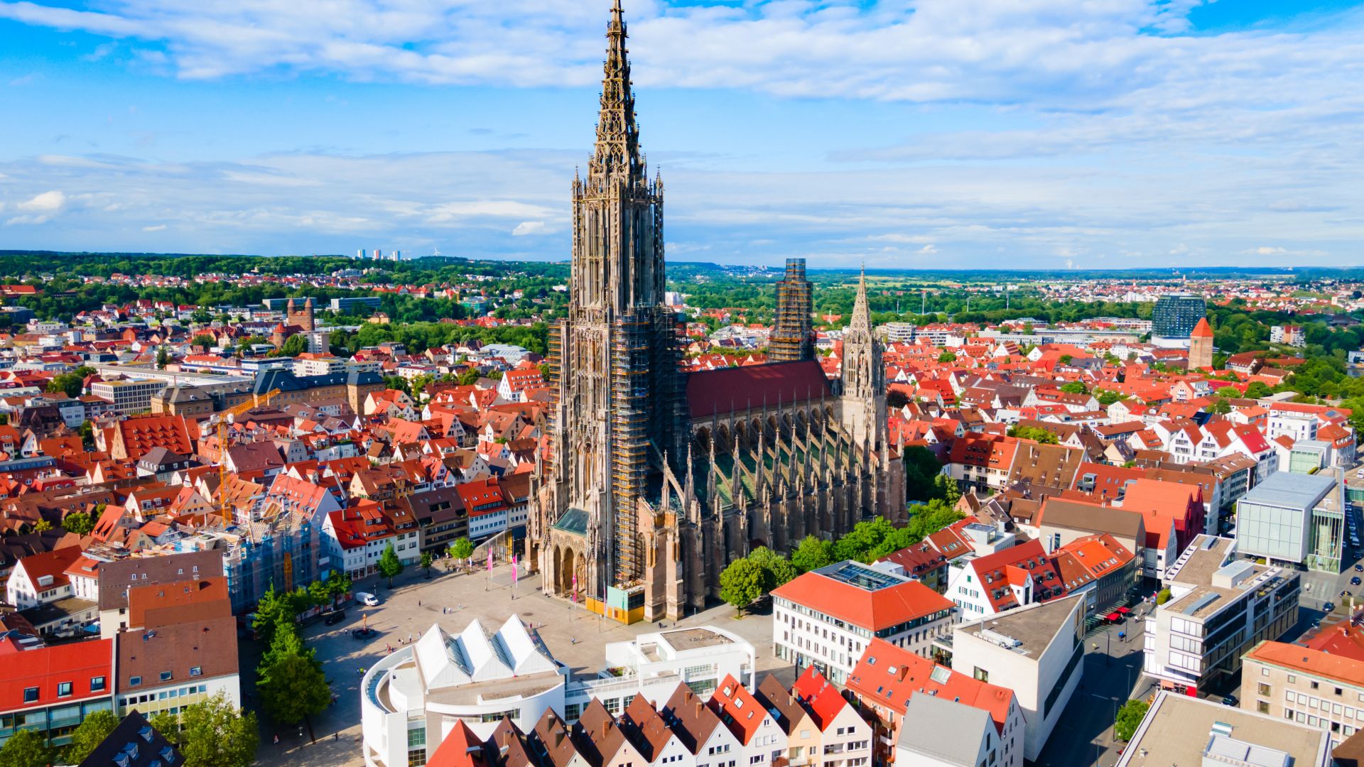 Aerial view of Ulm with the large Gothic Ulm Minster in the center of the picture. The surrounding area consists of numerous red rooftop houses and more modern buildings. A green landscape stretches out in the background.
