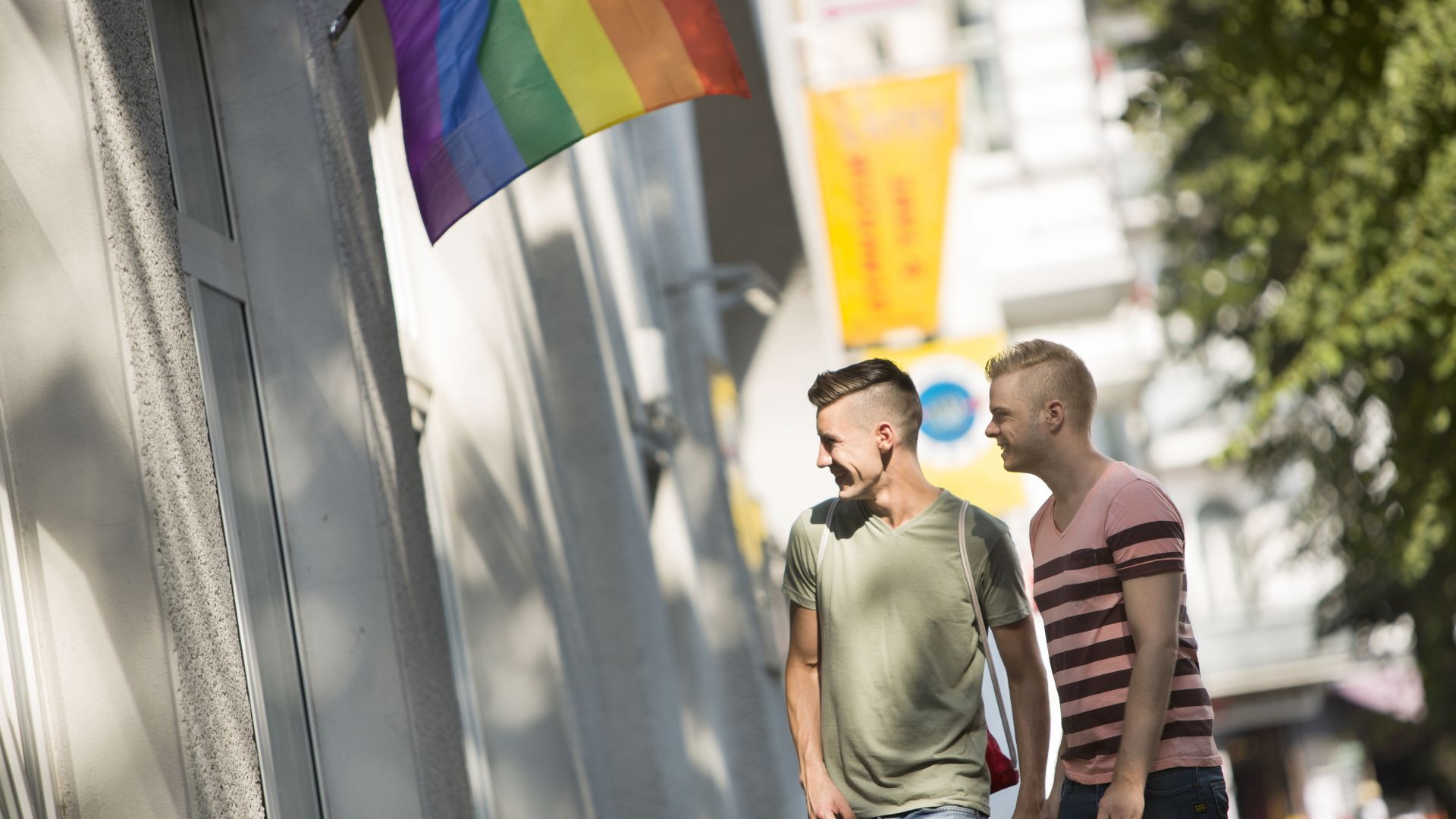 Zwei Männer gehen draußen neben einem Gebäude in Berlin spazieren. An dem Gebäude hängt eine Regenbogenflagge.